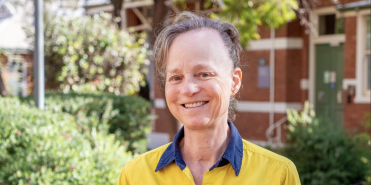 Student geologist Yeeda stand smiling on campus in front of a brick building and greenery. She wears her work uniform with her hair tied back.